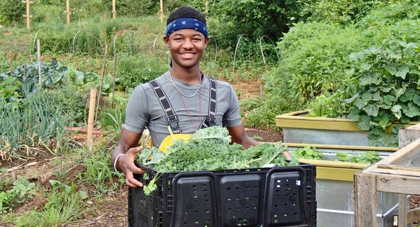 A person smiles at the camera while carrying a box filled with vegetables. A garden is in the background.